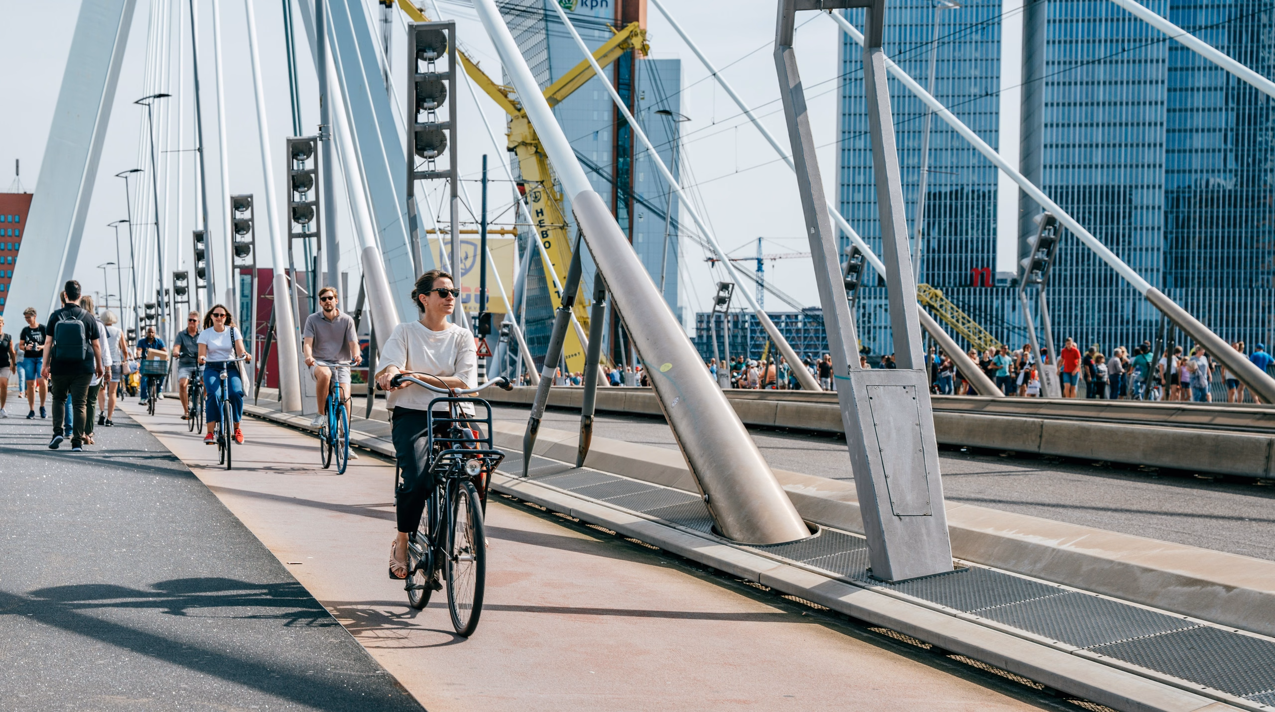 Cyclists and pedestrians crossing a bridge in a city with tall buildings behind them.
